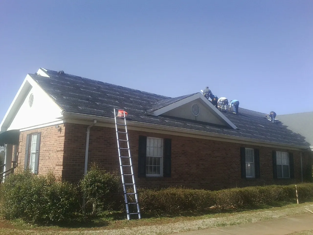 Crew tearing shingles off a brick ranch roof with a ladder set — Metal Roof Repair work by our crew in Warner