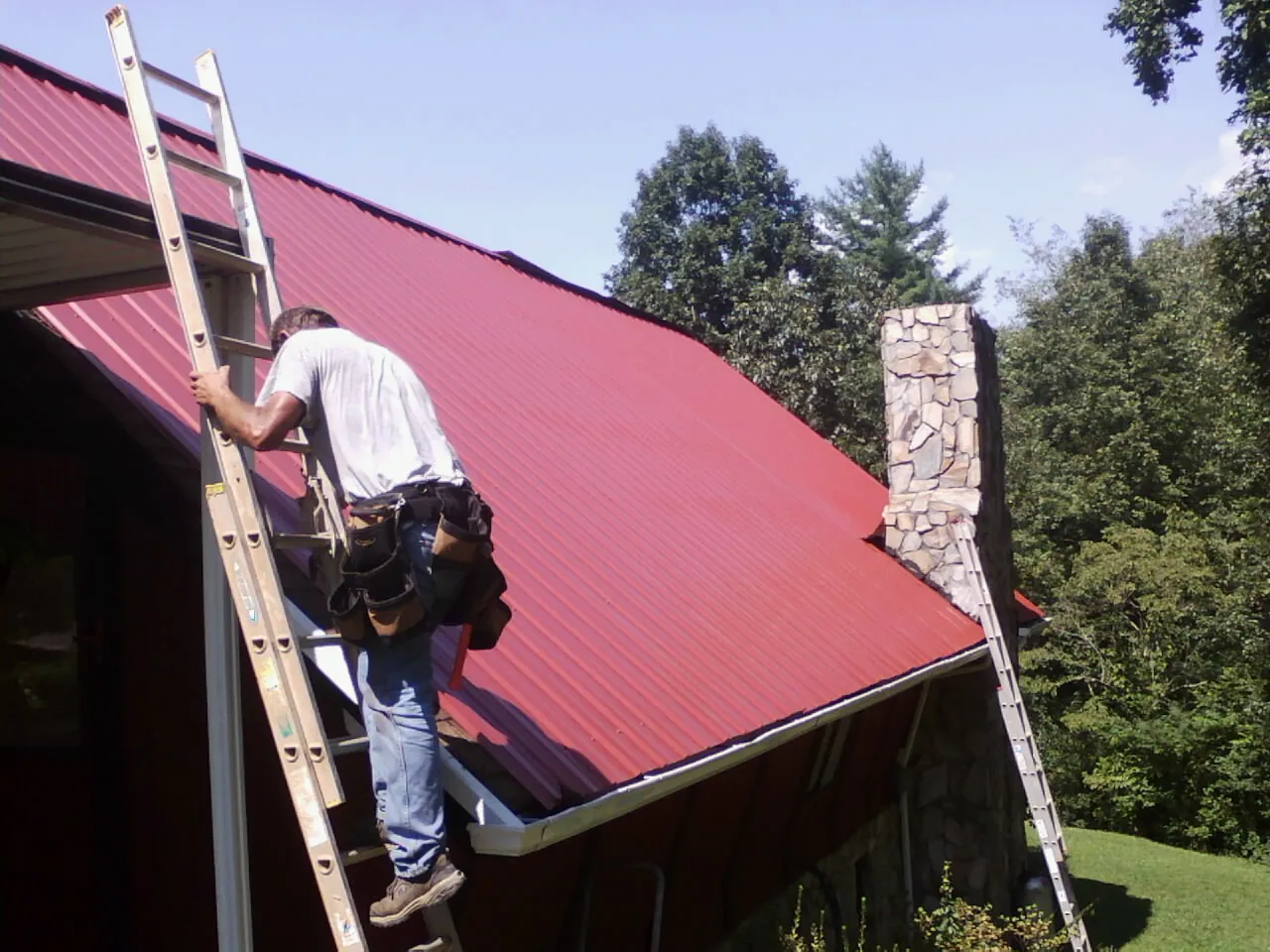 Roofer on a ladder beside a red metal roof and stone chimney — Soffit & Fascia Repair by Roofing Brothers in Warner